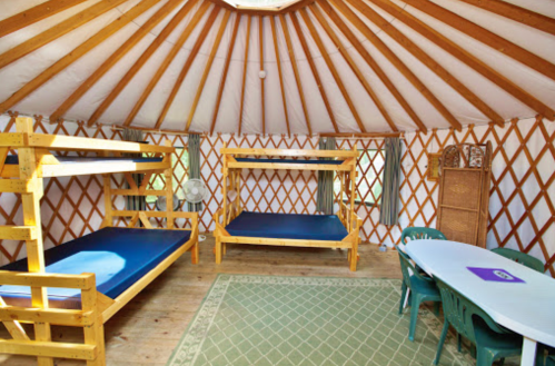 Interior of a yurt featuring bunk beds, a table with chairs, and wooden beams, with natural light coming through the ceiling.